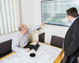 Besprechung vor Bauplan, Mitarbeiter zeigt auf Bildschirm